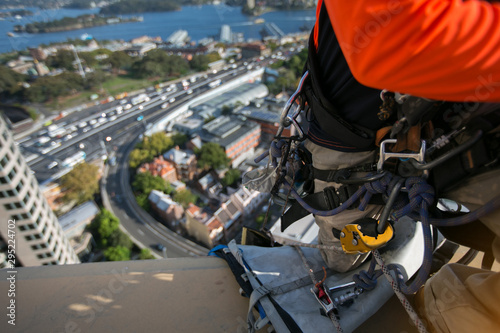 Abseiler wearing full body safety harness working at height hanging on the edge with descender clipping into the rope back up device ready to abseiling from high rise building road traffic background