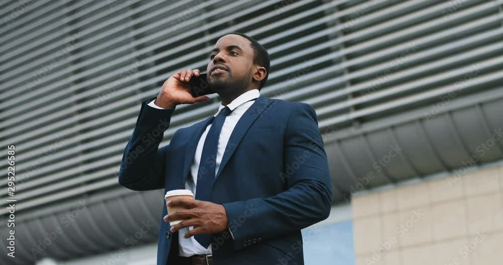 Handsome young African American businessman in suit and tie talking on the phone with a coffee to-go in hands while drinking it and standing outside in the morning before work. Handsome young African American businessman in suit and tie talking on the phone with a coffee to-go in hands while drinking it and standing outside in the morning before work.