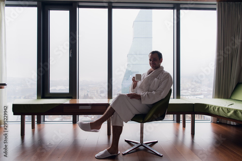 Young handsome carefree man wearing bathrobe near modern full length window enjoying a cup of coffee while looking outside, good morning. Horizontal photo banner for website header design with copy