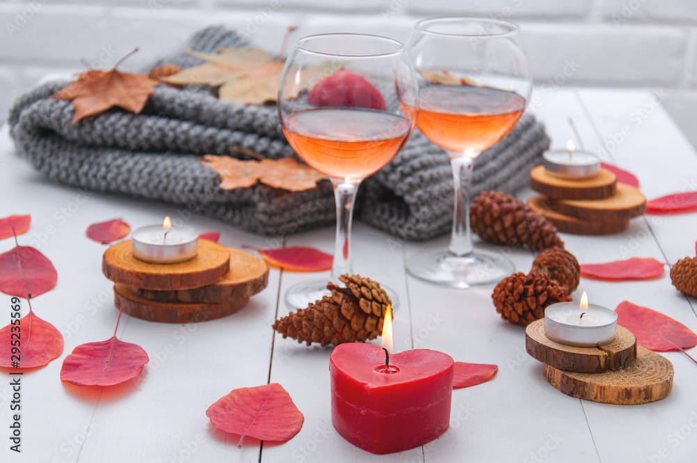 Small burning candles, two glasses with rose wine, cones, dry red leaves, a gray scarf knitted on a white wooden table. Hello, Autumn. Cozy autumn background.