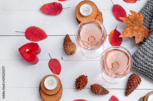 Small burning candles, two glasses with rose wine, cones, dry red leaves, a gray scarf knitted on a white wooden table. Hello, Autumn. Cozy autumn background.