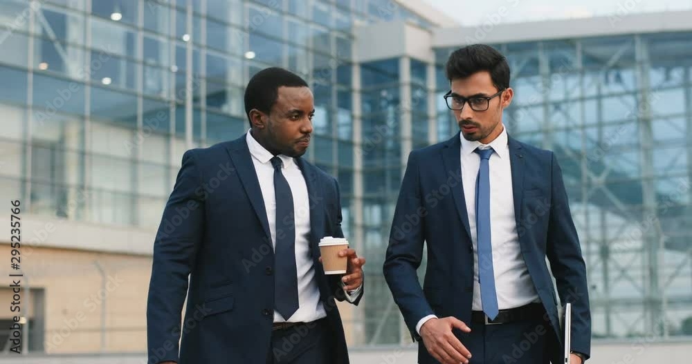 Two busy multiethnic handsoem men, business partners walking the office center city, drinking coffee to-go and talking about their plans.