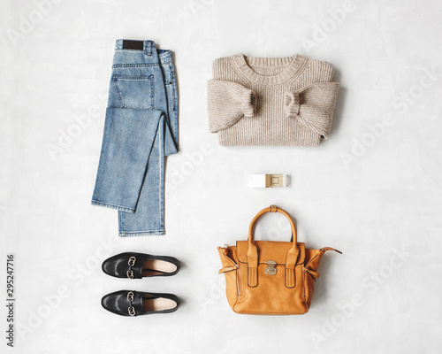 Blue jeans, beige knitted sweater, bag, black loafers or flat shoes on grey background. Overhead view of women's casual day outfit. Flat lay, top view. Women clothes.