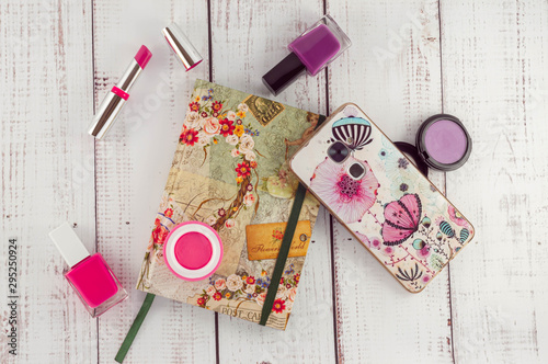 Several items for beauty, lipstick, nail polish, blush, eyeshadow, with a notebook and a smartphone on a wooden background