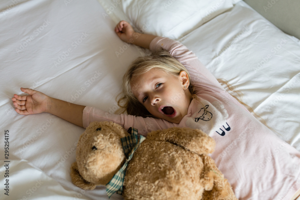 Top view of cute blonde little girl in pajamas and blindfold lying in white bed with teddy bear, awaking early in the morning before going to kindergarten. Bedtime, awakening and relaxation concept Top view of cute blonde little girl in pajamas and blindfold lying in white bed with teddy bear, awaking early in the morning before going to kindergarten. Bedtime, awakening and relaxation concept