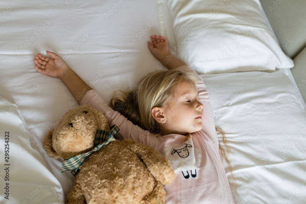 Top view of cute blonde little girl in pajamas lying in white bed with teddy bear, awaking early in the morning before going to kindergarten. Bedtime, awakening and relaxation concept