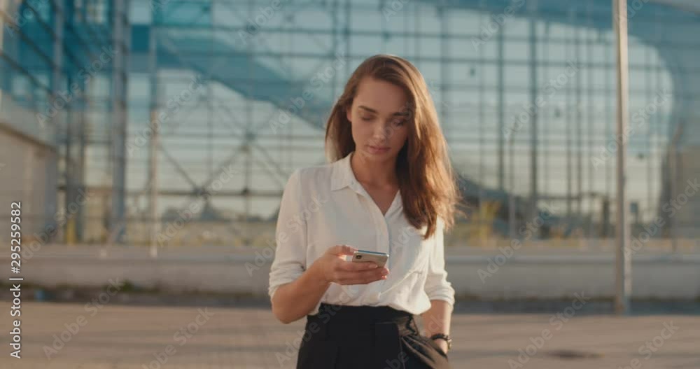 Attractive young Caucasian woman standing outdoor at the big modern glass building background, tapping and texting on the smartphone. Attractive young Caucasian woman standing outdoor at the big modern glass building background, tapping and texting on the smartphone.