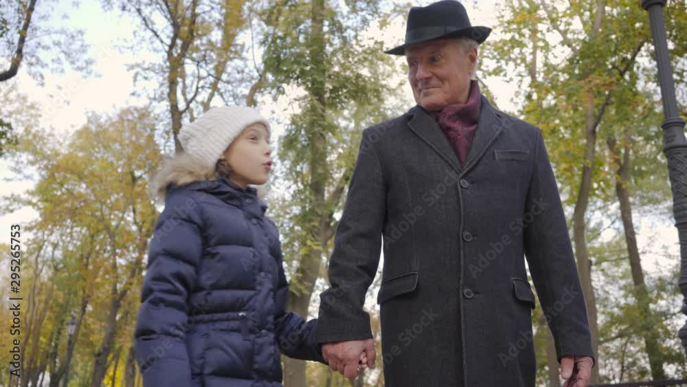 Pretty Caucasian girl in white warm hat spending weekends with her grandfather. Child jumping around senior smiling man in elegant black hat and coat. Pretty Caucasian girl in white warm hat spending weekends with her grandfather. Child jumping around senior smiling man in elegant black hat and coat.