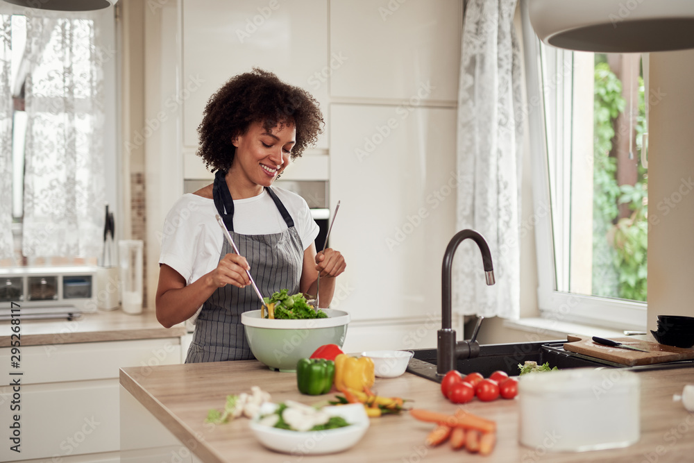 Attractive mixed race woman in apron mixing vegetables in bowl while standing in kitchen at home. Attractive mixed race woman in apron mixing vegetables in bowl while standing in kitchen at home.