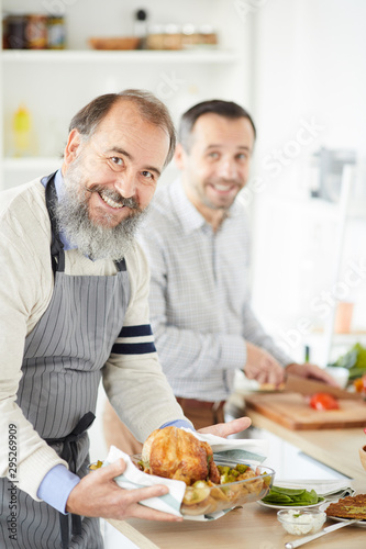 Portrait of senior man with beard smiling at camera while preparing roast turkey with young man helping him in the background