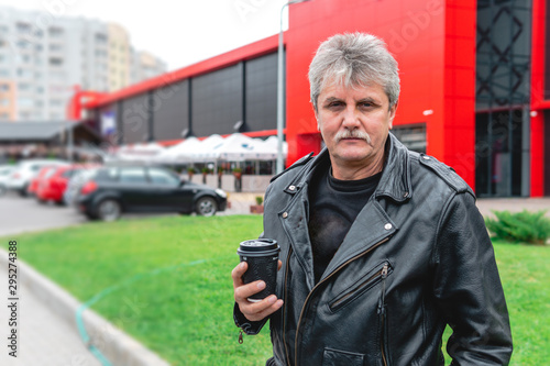 middle-aged man holding a cup of coffee, brutal biker style in a leather jacket. coffee to go, outdoor street. urban