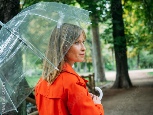 фотографія Caucasian girl with red gabardine walks through a park under a transparent umbrella