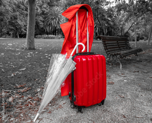 Photograph of a travel suitcase, an umbrella and a red gabardine in a park.