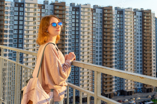 Tween redhead girl in pullover, jeans and sunglasses standing on balcony against high-rise multi-storey residential building at sunset. Beautiful look, fashionable city street outfit, teenage fashion