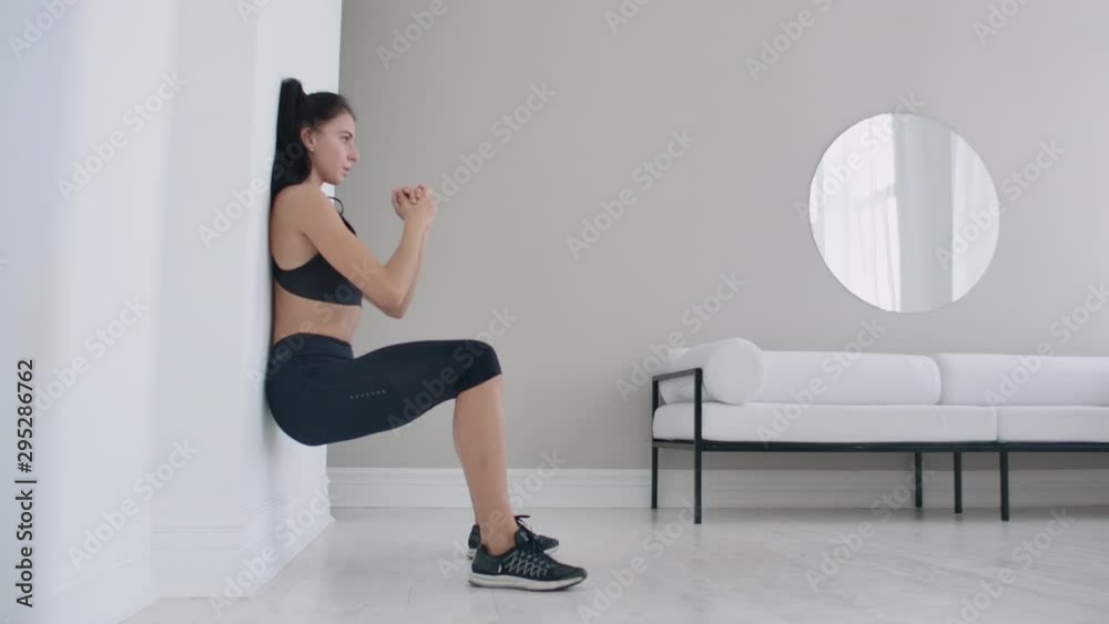 The brunette girl froze in a squat leaning on the wall doing a static exercise The brunette girl froze in a squat leaning on the wall doing a static exercise