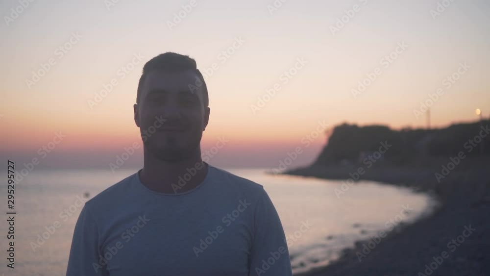 A young man stands on a background of sea sunset. A man is looking at the camera while standing on the beach. A man stands in a sea sunset on the coast. A young man stands on a background of sea sunset. A man is looking at the camera while standing on the beach. A man stands in a sea sunset on the coast.