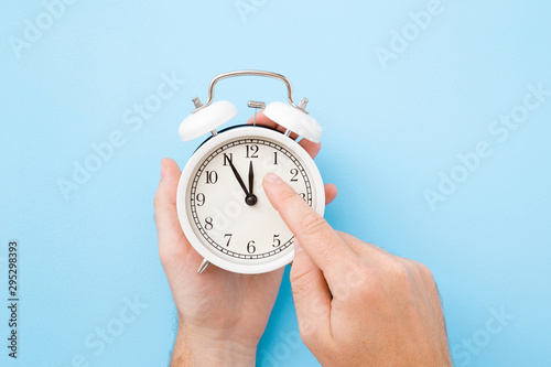 Man hand holding white alarm clock. Finger pointing to arrow of twelve o'clock. Light pastel blue background. Time change concept. Closeup.