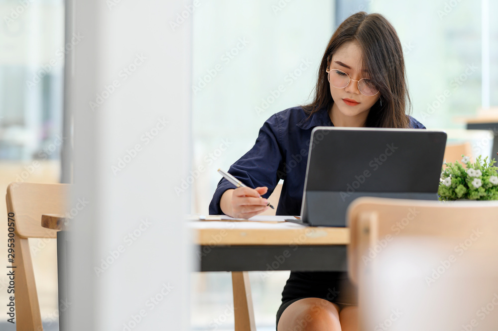 Young woman holding pen and working with laptop while sitting in co-workspace. Young woman holding pen and working with laptop while sitting in co-workspace.