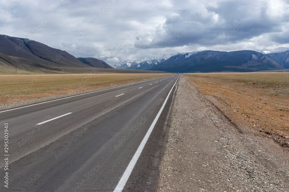 The gray asphalt road goes far and abuts against blue-blue clouds and mountains on a cloudy autumn day in the Altai Mountains