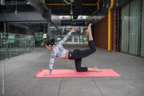 Young attractive smiling woman practicing yoga, sitting in One Legged King Pigeon exercise, Eka Pada Rajakapotasana pose, working out, wearing sportswear, grey pants, bra, indoor full length, home