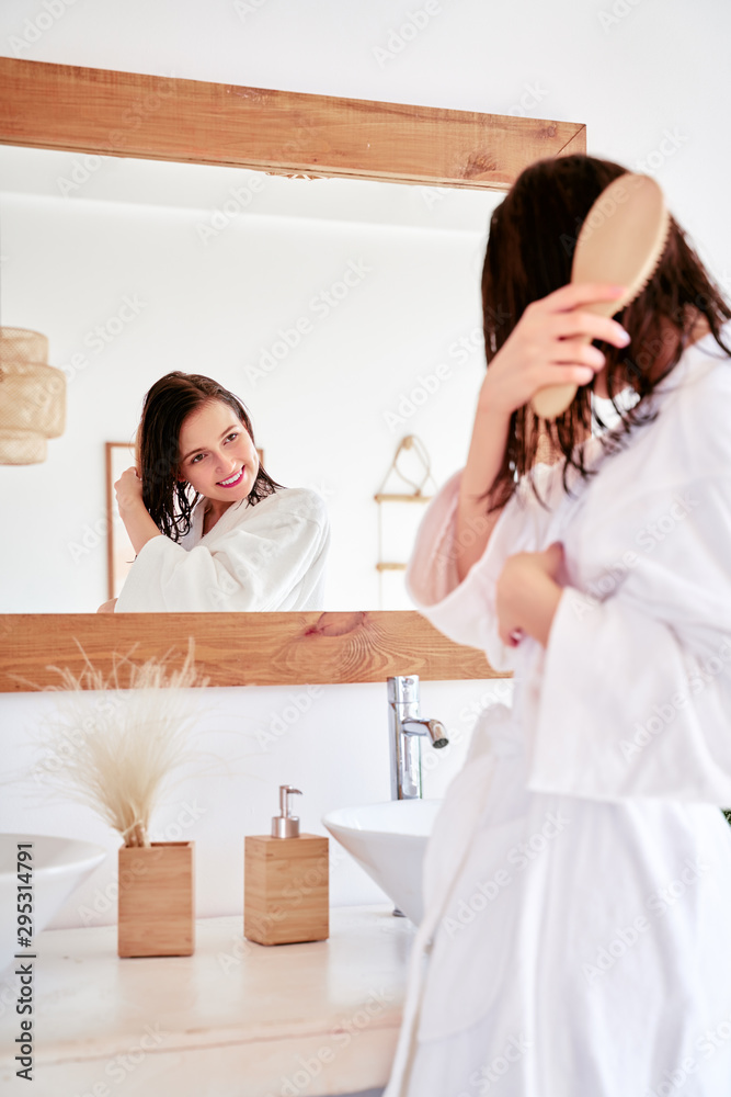 Photo of brunette combing her hair in front of bathroom mirror Photo of brunette combing her hair in front of bathroom mirror