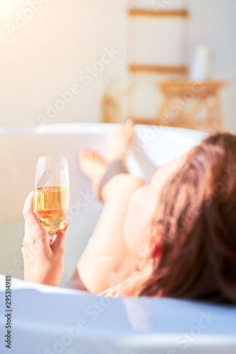 Photo from back of young woman with wine glass with wine in her hands lying in bath in apartment.