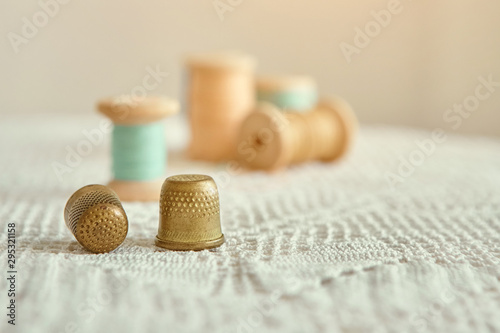 Two vintage thimbles close-up, in the background old wooden bobbins with sewing threads. Are on the tablecloth antique hand knit lace.