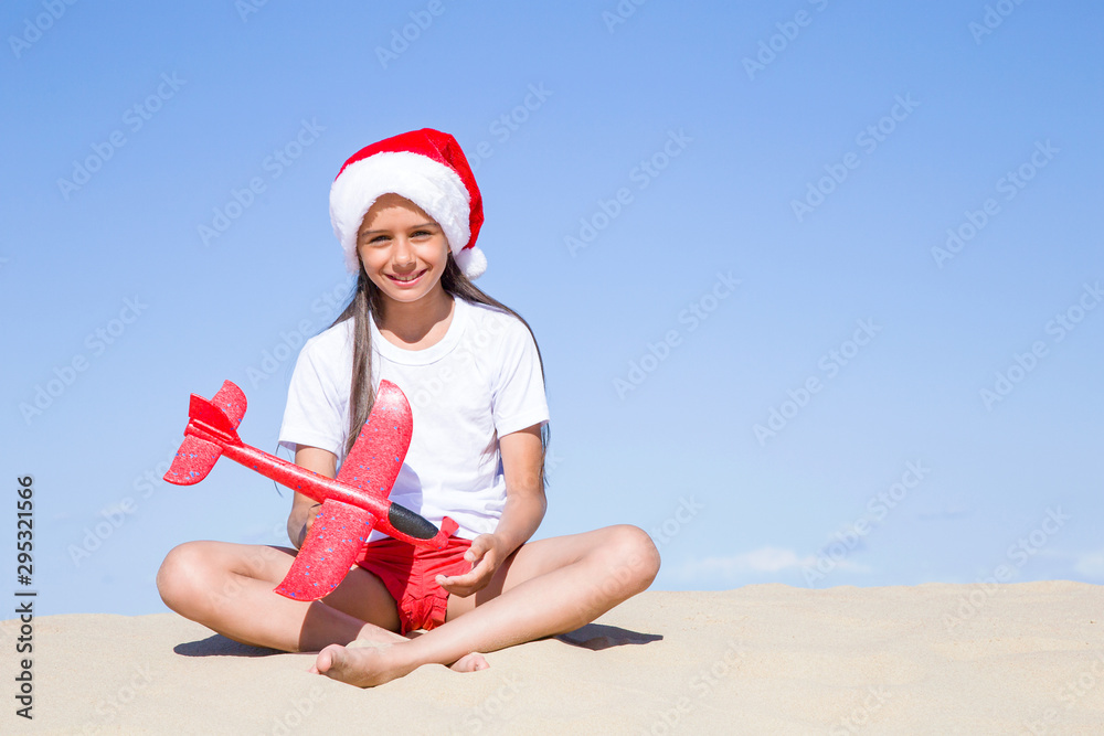 Obraz premium Christmas celebration vacations on a tropical beach: Happy cute little girl wearing a red Santa hat sitting on the sandy beach by the sea and holding a red toy plane on clear sunny day