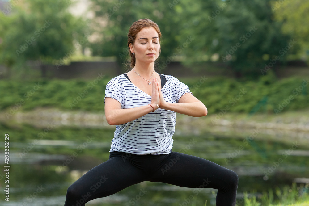 Young beautiful woman doing yoga exercise in green park near the pond. Exercices for improve the flexibility. Wellbeing and wellness. Healthy lifestyle and fitness concept.