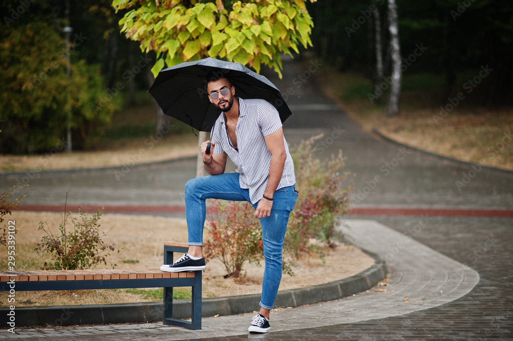 Fashionable tall arab beard man wear on shirt, jeans and sunglasses with umbrella posed at rain on park square.