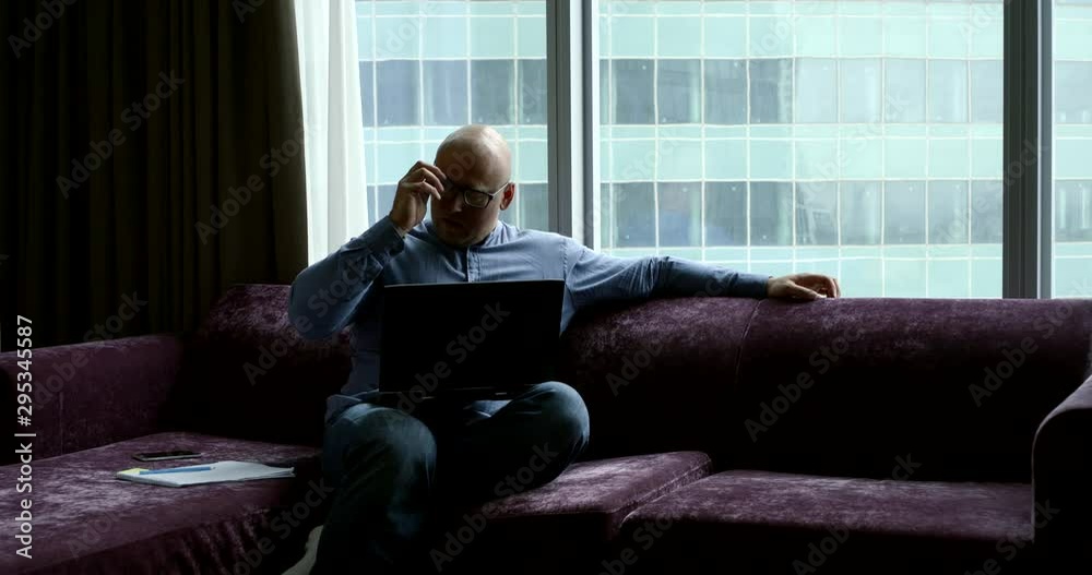 bald man sitting on the couch by the window and working on a laptop. he takes off his glasses, rubs his face, picks up the phone bald man sitting on the couch by the window and working on a laptop. he takes off his glasses, rubs his face, picks up the phone