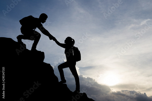 Silhouette of people helping each other hike up a mountain at sunset background. Teamwork, success and goal concept.