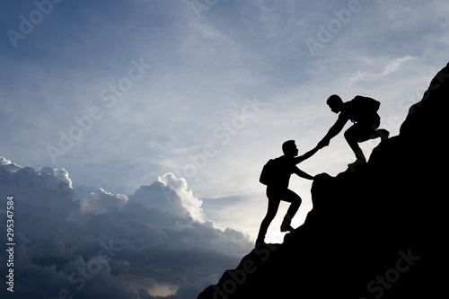 Silhouette of people helping each other hike up a mountain at sunset background. Teamwork, success and goal concept.