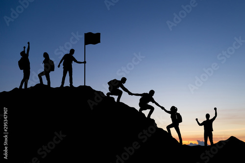 Silhouette of people helping each other hike up a mountain at sunset background. Teamwork, success and goal concept.