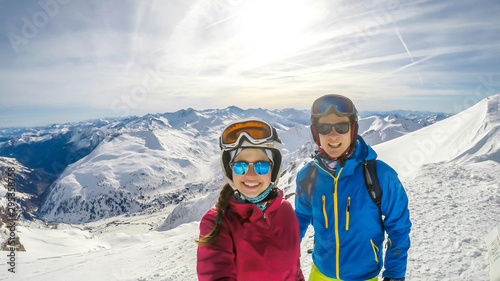 A couple in skiing outfits standing on top of a snowy mountain peak and enjoying the sunny day. Both of them are laughing and enjoying the view. Tall Alpine peaks all around the couple. Togetherness.