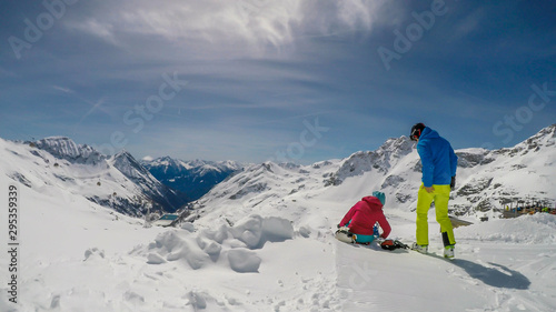A couple in skiing outfits, sitting on the snow and enjoying the snowy mountains. Endless ranges of snow caped mountains in front of them. Togetherness, enjoying the time. Winder paradise.