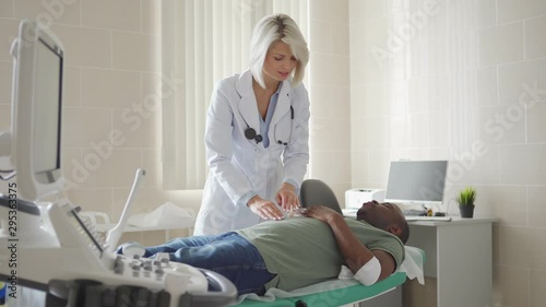 Tracking shot of female physician in white coat palpating belly of young African patient with bandage on arm after venipuncture lying on couch in doctors office
