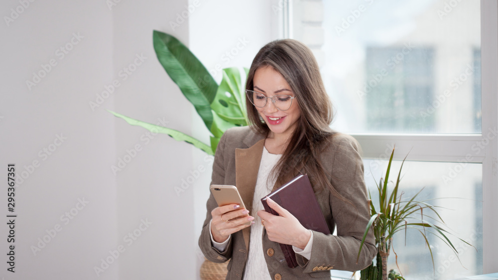 Young brunette woman uses the phone standing at the window in a bright comfortable office. Young brunette woman uses the phone standing at the window in a bright comfortable office.