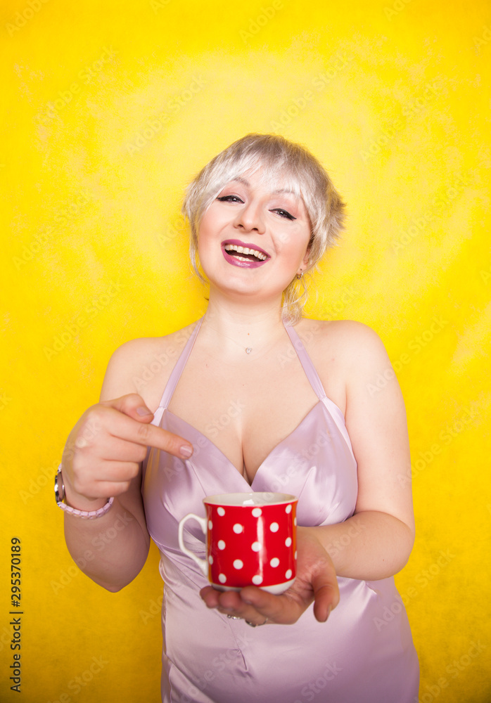 pretty curvy young blonde woman with short hair enjoys drinking tea from cute red polka dot ceramic cup on yellow studio background pretty curvy young blonde woman with short hair enjoys drinking tea from cute red polka dot ceramic cup on yellow studio background