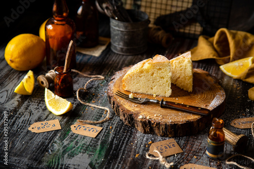 homemade two tasty slices of baked lemon biscuit cake with powdered sugar on top stands on wooden board on rustic table with lemons  old bottles opposite concrete wall  selective focus