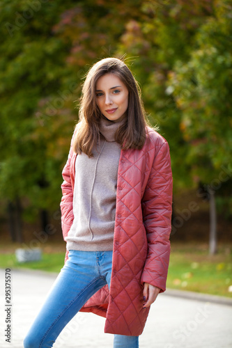 Young stylish woman in a salmon down jacket posing on a background of autumn park © Andrey_Arkusha