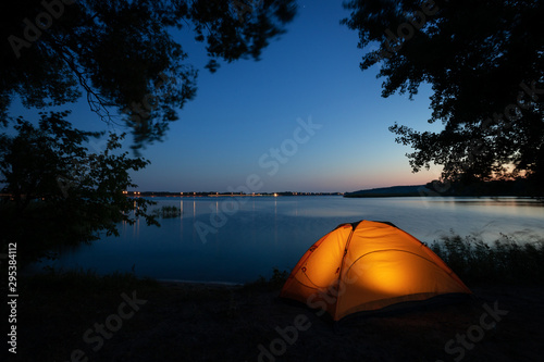 Orange illuminated inside tent surrounded by silhouette of tree branches on lake at night