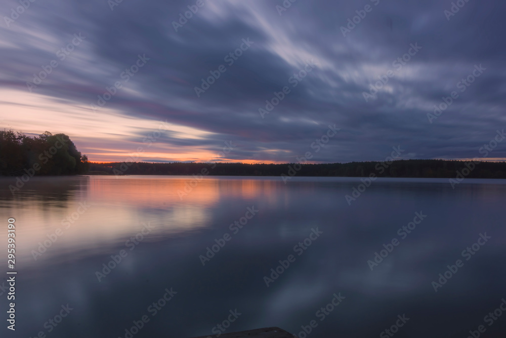 Landscape; lake and trees before sunrise, long exposure blurred objects, dark clouds and sky, autumn trees and orange sun are reflected in the water