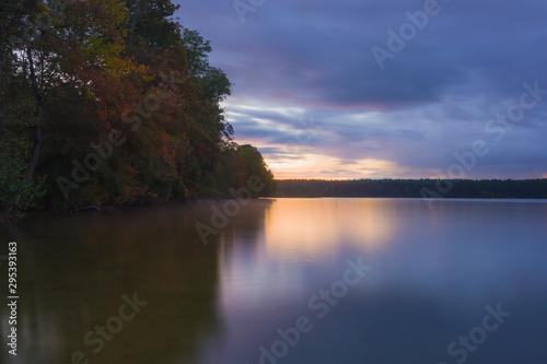 Landscape; lake and trees before sunrise, long exposure blurred objects, dark clouds and sky, autumn trees and orange sun are reflected in the water