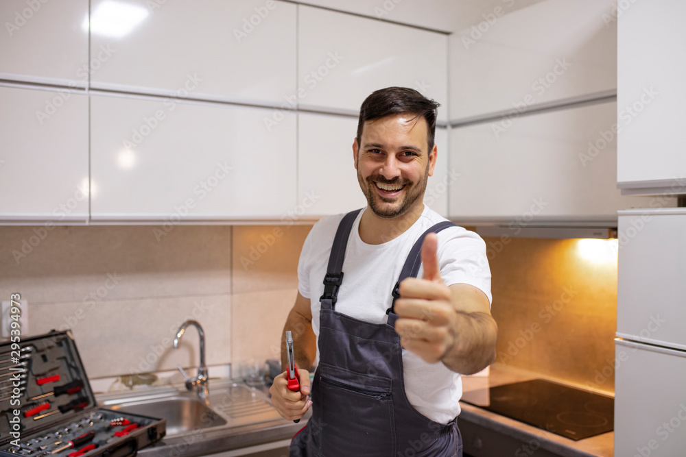Portrait of a handyman in uniform standing in the renovated kitchen at home showing thumb up. Young worker with tool bag standing near oven in kitchen Portrait of a handyman in uniform standing in the renovated kitchen at home showing thumb up. Young worker with tool bag standing near oven in kitchen