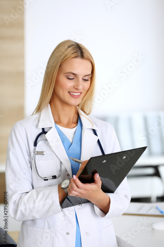 Doctor woman at work in hospital excited and happy of her profession. Blonde physician controls medication history records and exam results while using clipboard. Medicine and healthcare concept