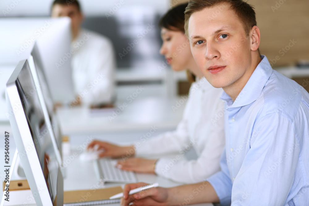 Obraz premium Cheerful smiling businessman headshot at work in modern office. Unknown casual dressed entrepreneur using pc computer while sitting with diverse colleagues at the background. Multi ethnic working