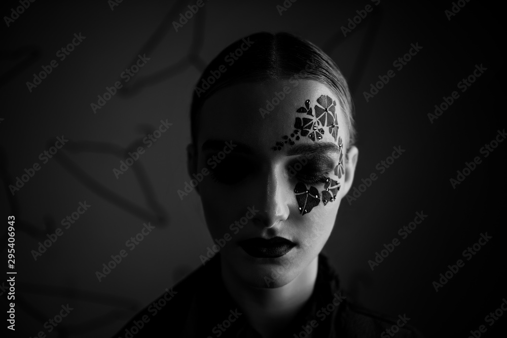 Close-up portrait of a girl with clean skin, bright professional make-up, purple lipstick, face with black ivy tattoo around her eyes with rhinestones. Gray background, pattern of hangers. Fashion