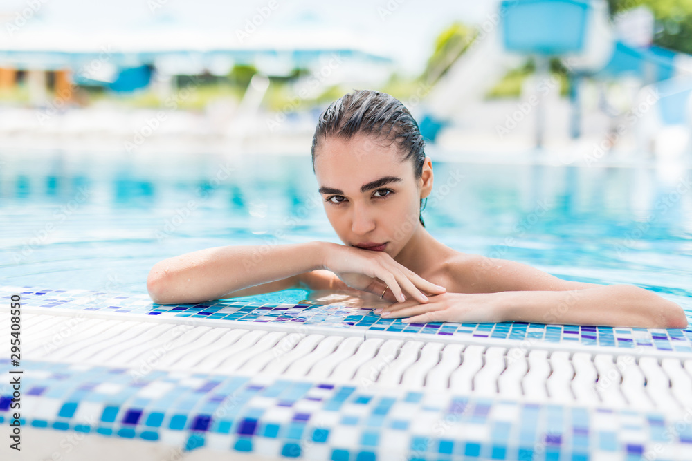 Portrait of attractive girl relaxing in swimming pool, posing near edge. Beautiful young woman looking at camera with toothy smile at poolside, having rest at spa resort in summer.