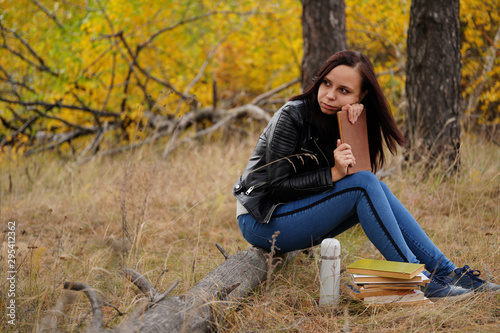 A young beautiful woman with long dark hair in casual clothes is sitting on a log, holding a book in her hands, and thoughtfully looking to the side.